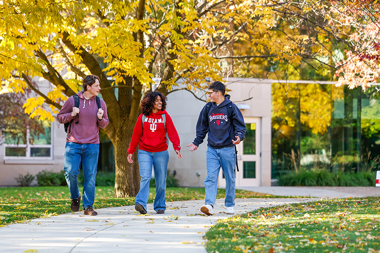 two male and one female student walking on the IU South Bend campus in the fall, trees are yellow and leaves have fallen along the path as they walk toward the foreground.
